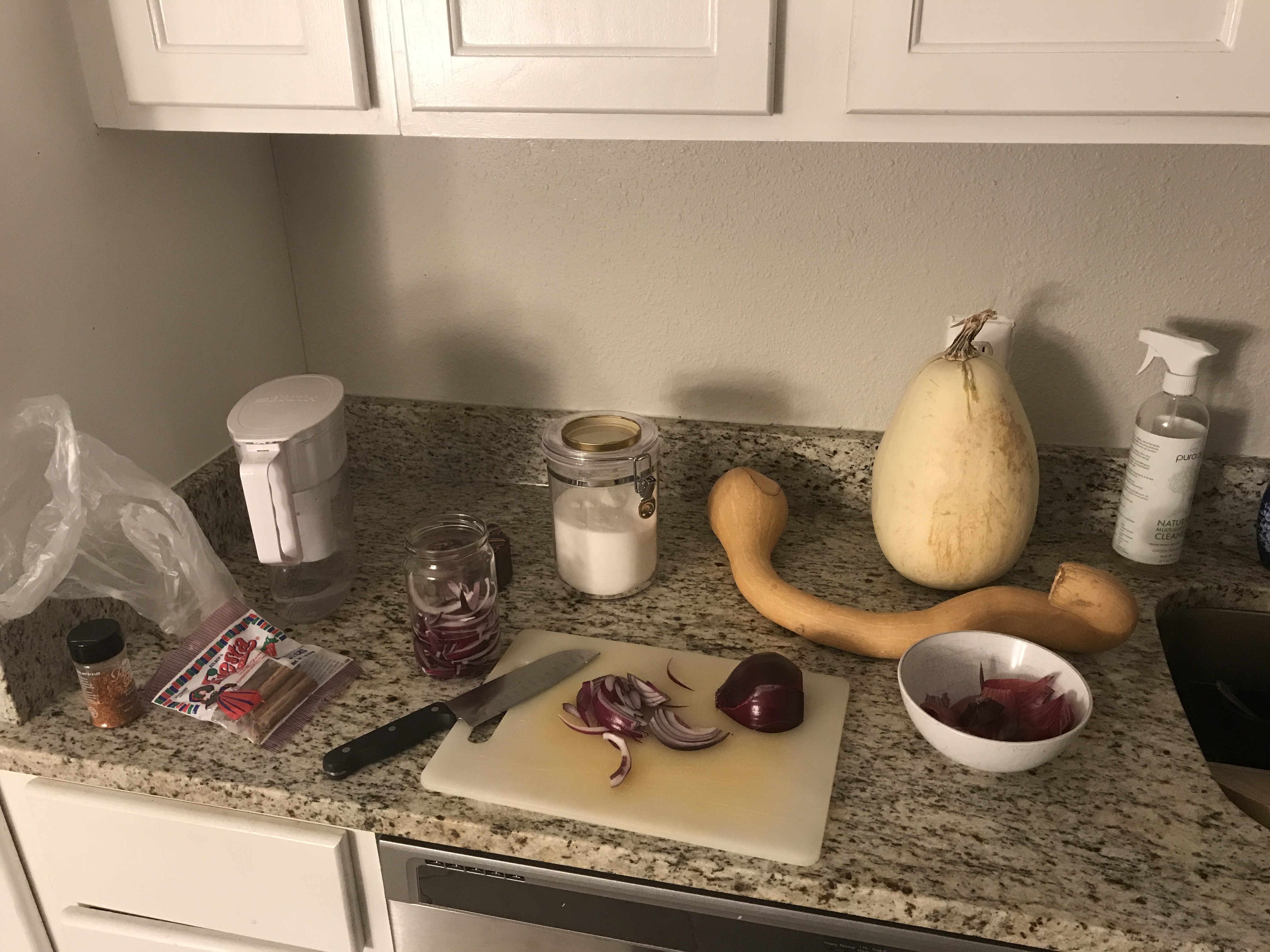 A messy kitchen counter containing a cutting board with a red onion, a couple of weird pumpkins that I avoided cooking into pie, and a half-full jar for fermenting in.